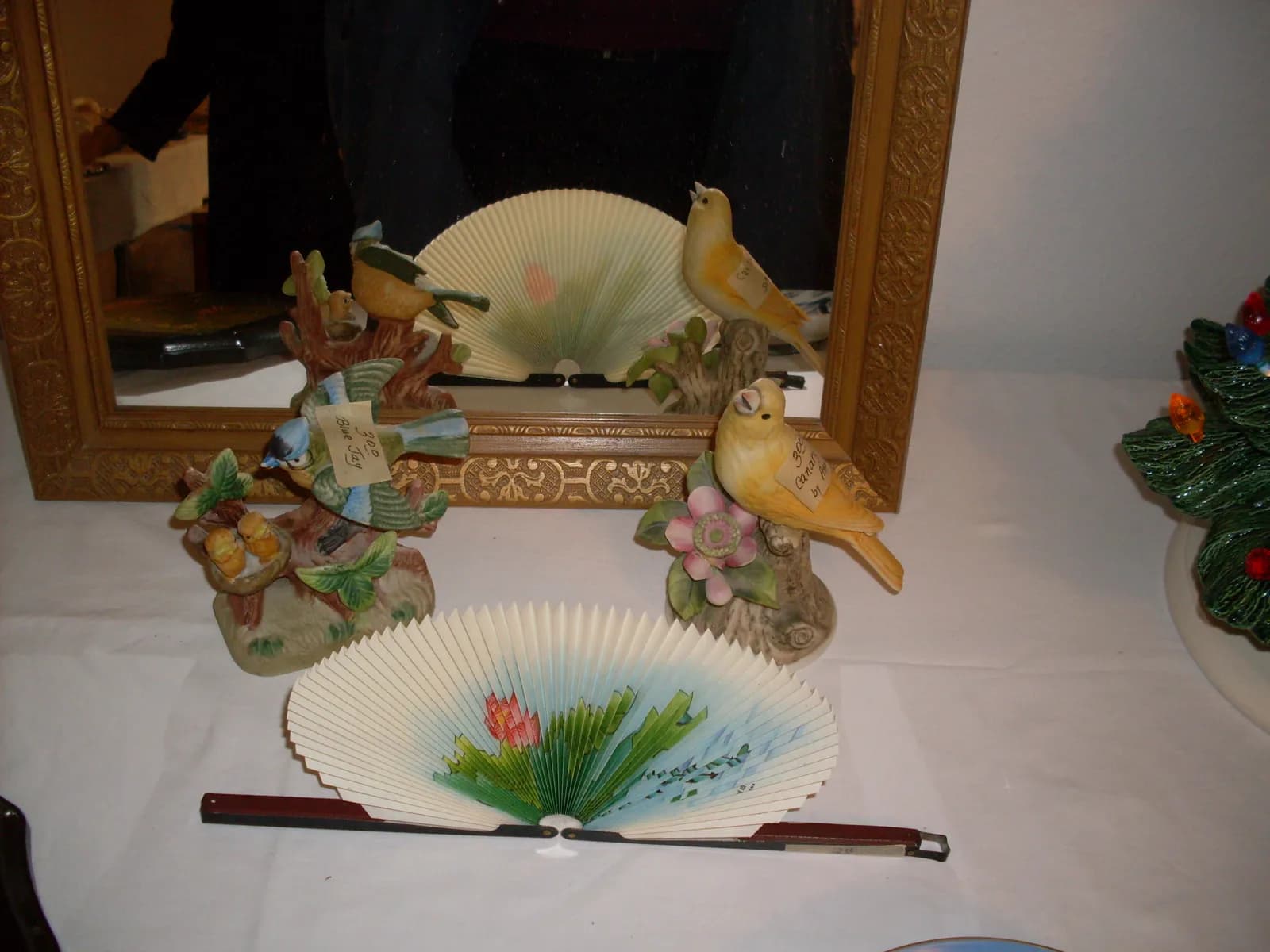 Porcelain bird figurines with hand-written price tags beside a painted paper fan, in front of a gilt-framed picture.