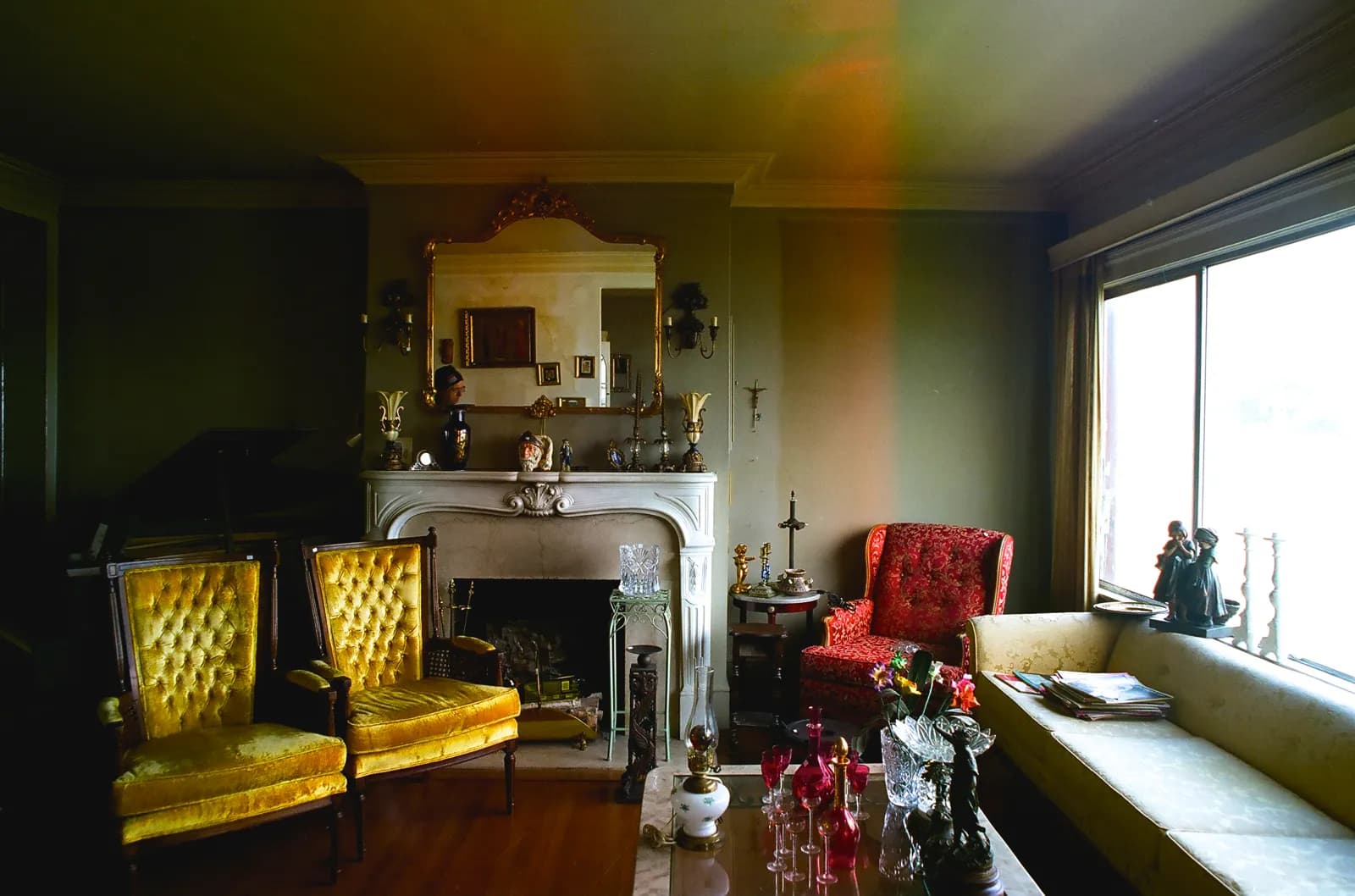 A living room set out for an estate sale: two gold tufted velvet armchairs, a red brocade wingback, a cream damask sofa and a marble fireplace with a gilt mirror.