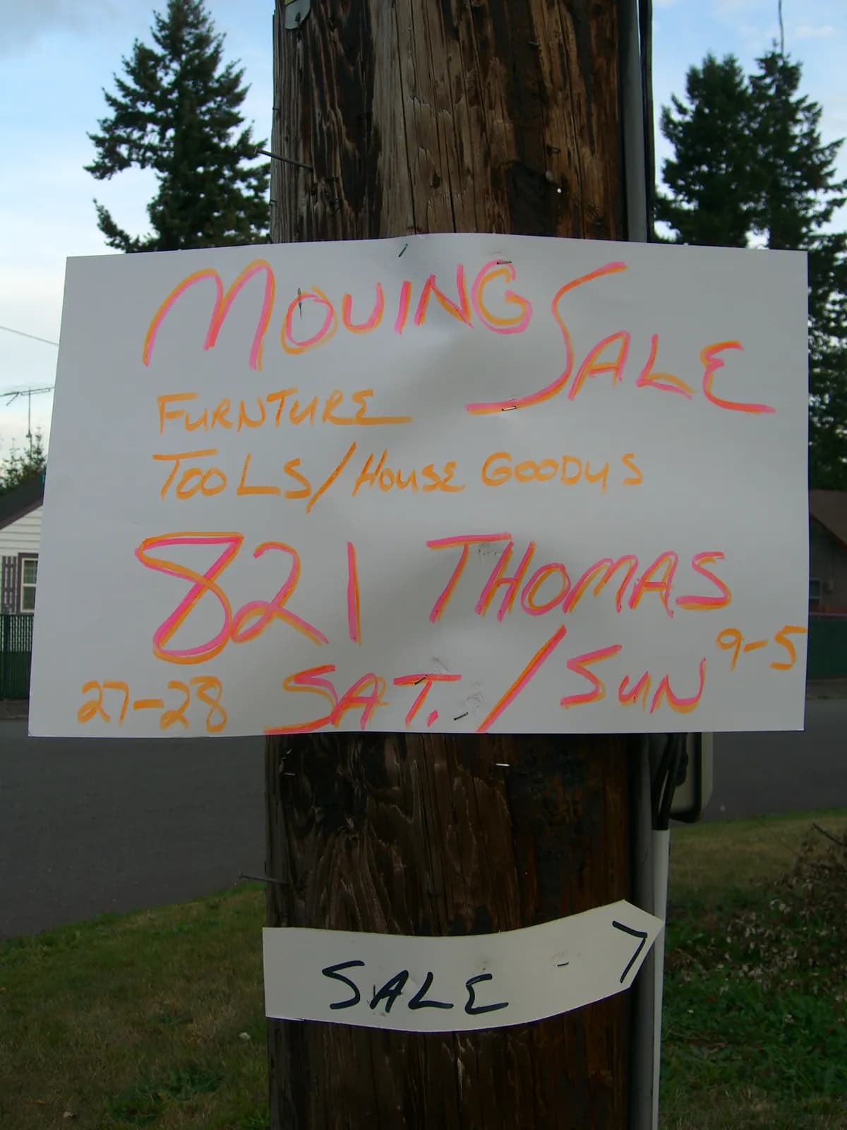 A hand-lettered Moving Sale poster in pink and orange marker stapled to a wooden utility pole, listing furniture, tools and house goods.