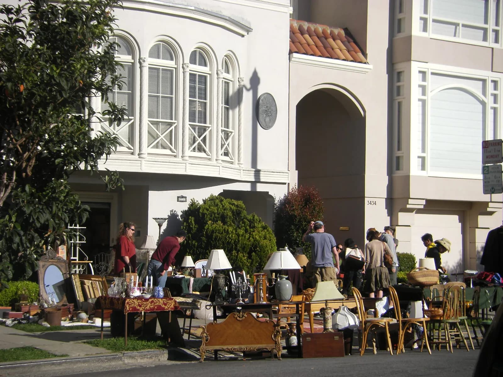 A garage sale on the sidewalk in front of cream stucco row houses, with lamps, candlesticks, a carved headboard and chairs.