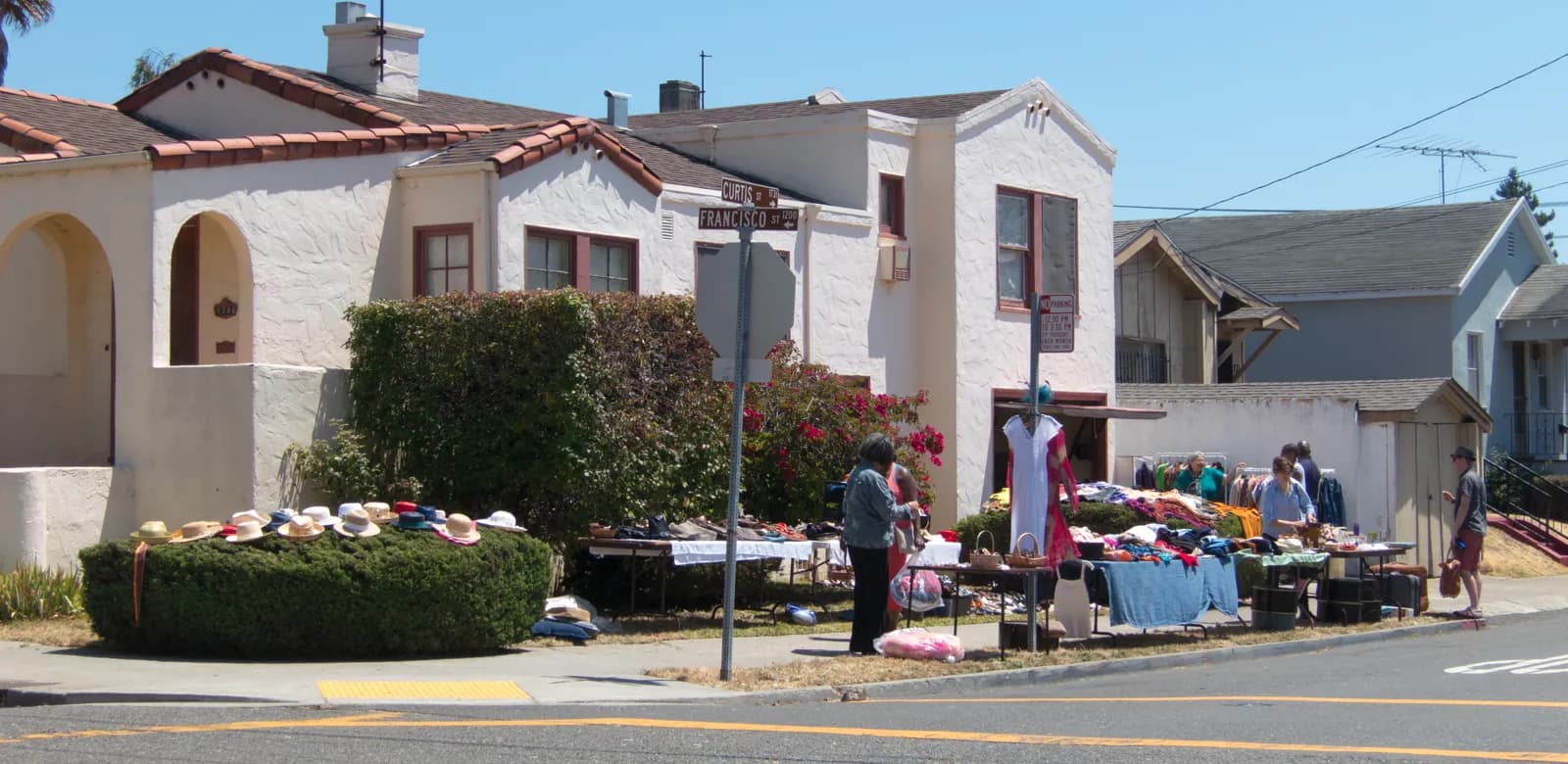 A corner yard sale outside white stucco Spanish-style houses with red tile roofs: straw hats laid out on a clipped hedge and tables of shoes and clothes.