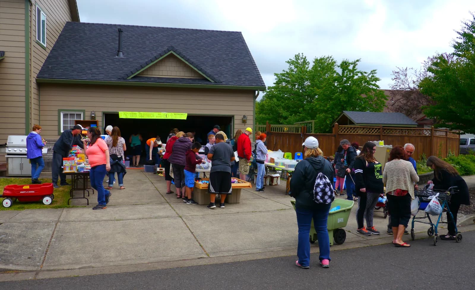 Shoppers crowd a driveway and open garage at a neighborhood-wide garage sale, browsing tables of toys and kids' clothes under a yellow banner.