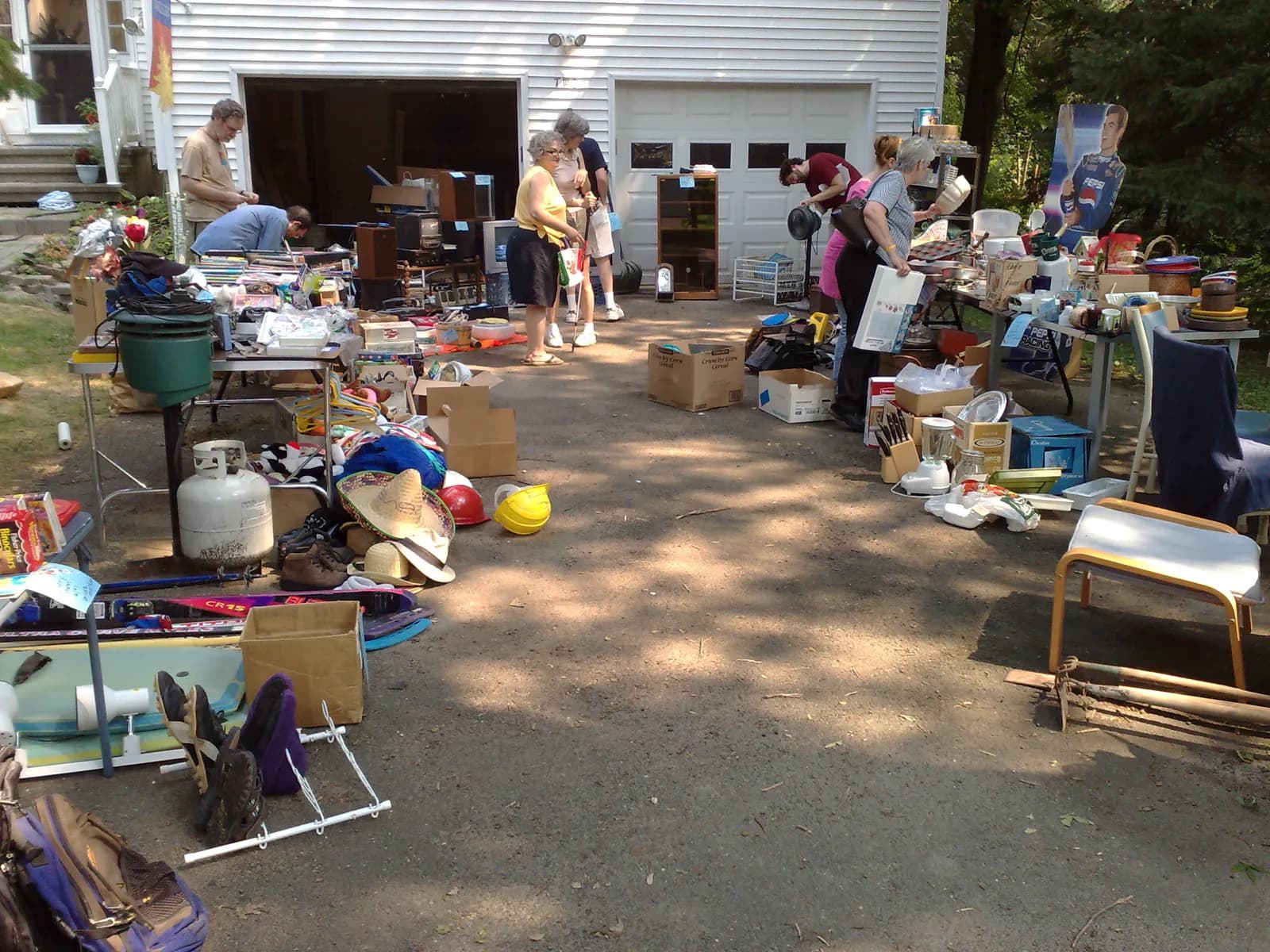 A moving sale filling a long driveway in front of a white two-car garage, with tables of kitchenware, electronics and books, boxes and skis.