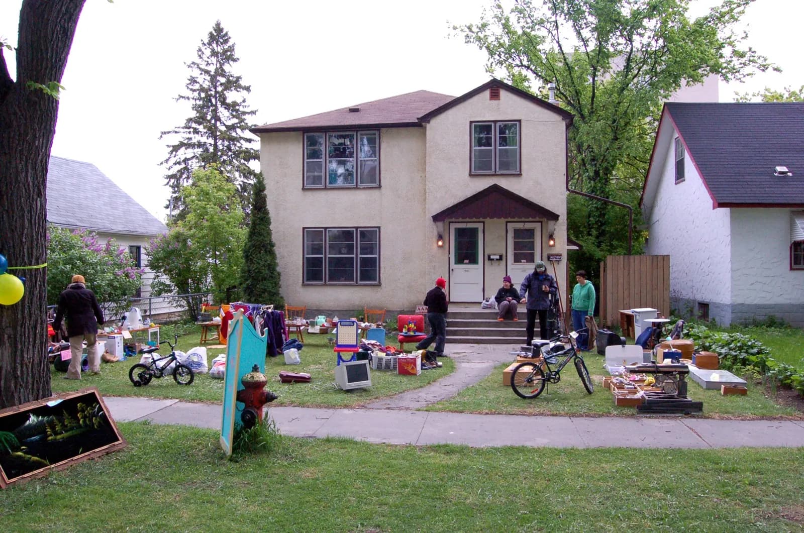 A stucco duplex seen from across the sidewalk with a yard sale on the front lawn: bikes, a computer monitor, chairs, a clothes rack and boxes of tools.