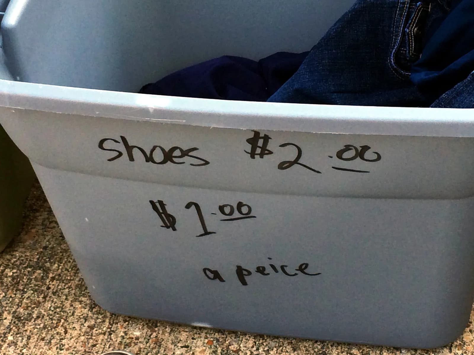 A pale blue plastic tub of clothes at a yard sale, with prices hand-written on its side in marker: shoes $2.00.