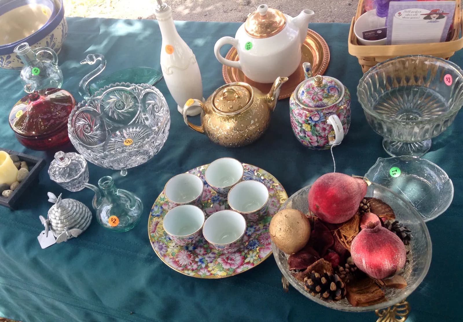 A yard sale table covered in a teal cloth with glassware and china, each piece marked with a round colored price sticker of $2, $3, $10 or $20.