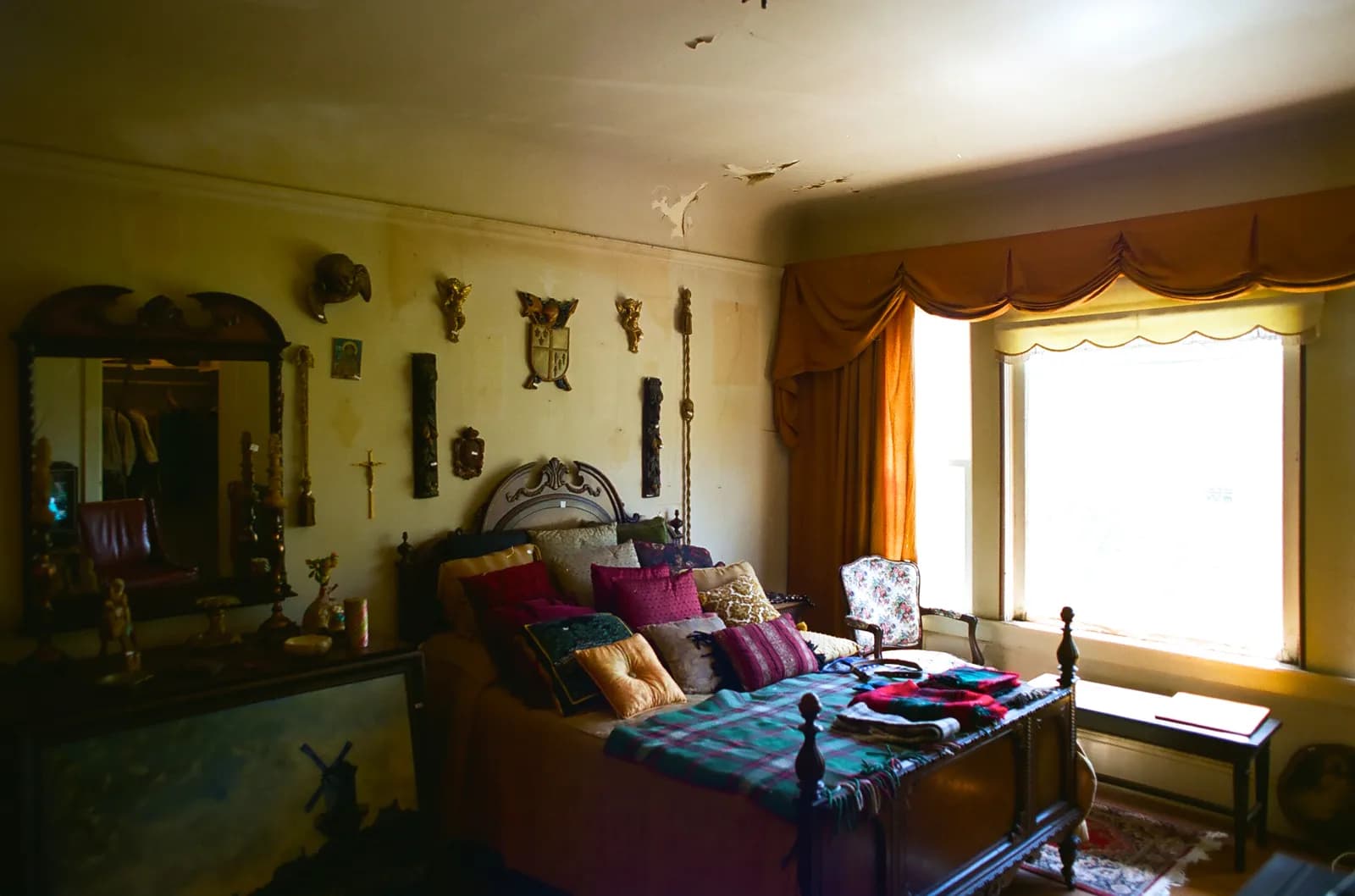 A bedroom during an estate sale: a carved wooden bed piled with cushions and folded plaid blankets, an ornate mirror, a dresser and carved wall pieces.