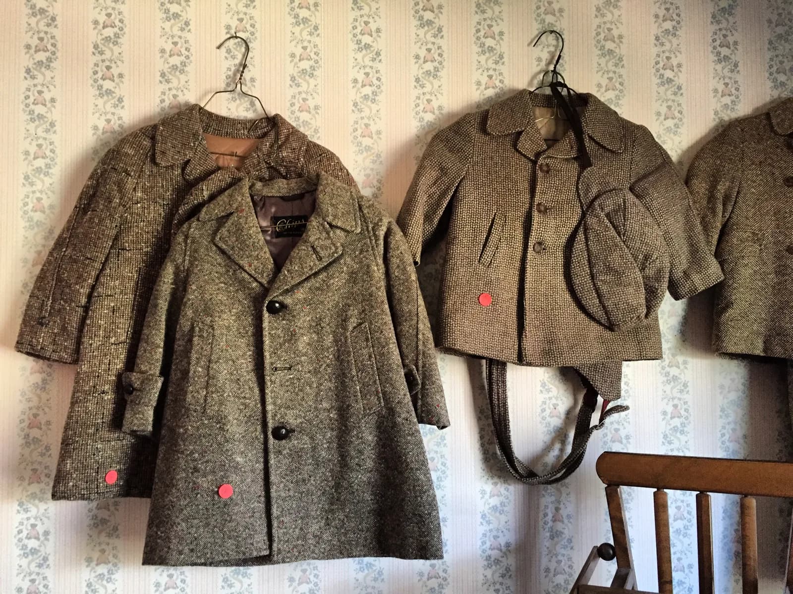 Vintage children's tweed coats, a jacket and a matching flat cap on wire hangers against floral wallpaper, each marked with a pink dot price sticker.