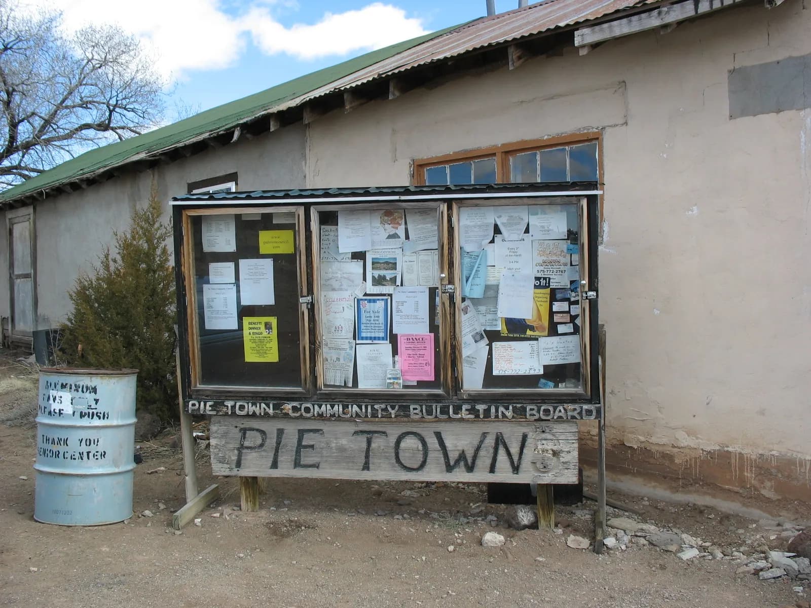A glass-fronted wooden bulletin board outside an adobe building, crowded with paper flyers including a For Sale notice.