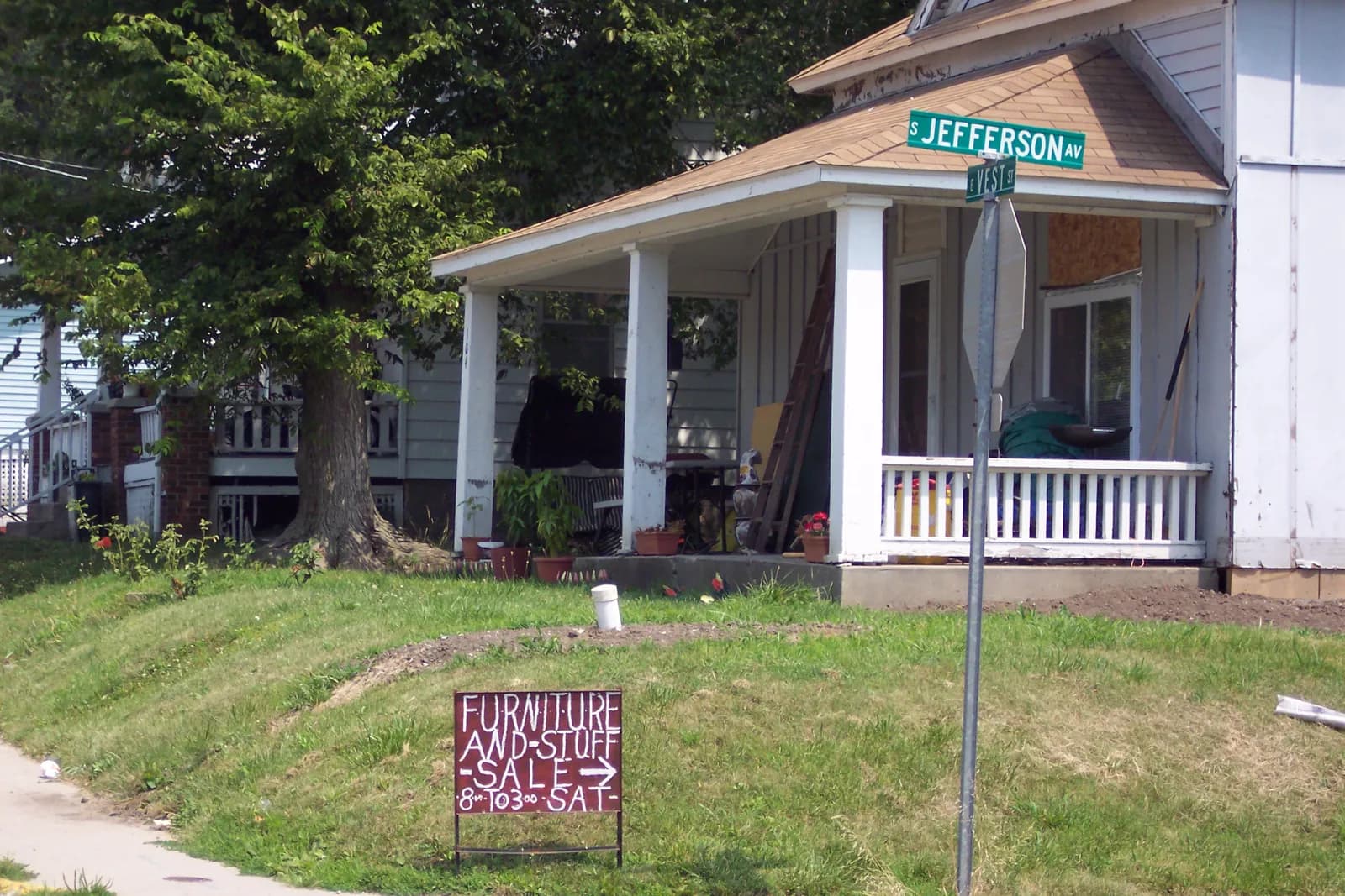 A hand-painted maroon sign reading Furniture and Stuff Sale, with an arrow and 8 to 3 Sat, standing on a grassy street corner below the street name signs.
